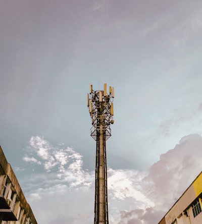 A tall communication tower stands prominently against a backdrop of a cloudy sky. The tower is equipped with multiple antennas and electronic equipment, suggesting its function in telecommunications. Surrounding the tower, there are parts of buildings visible in the lower corners of the image, indicating an urban setting.