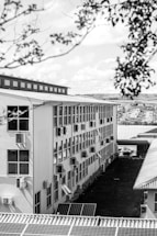A multi-story building with numerous windows and air conditioning units lining the walls. The structure has a corrugated rooftop, and solar panels are visible in the foreground. The scene includes surrounding greenery and trees, with a view extending to distant housing structures under a partly cloudy sky.