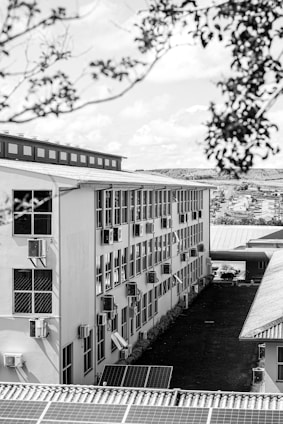 A multi-story building with numerous windows and air conditioning units lining the walls. The structure has a corrugated rooftop, and solar panels are visible in the foreground. The scene includes surrounding greenery and trees, with a view extending to distant housing structures under a partly cloudy sky.