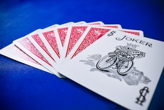 A set of playing cards spread out on a vibrant blue surface, highlighting a joker at the forefront. The backs of the other cards feature a red intricate design with white borders.