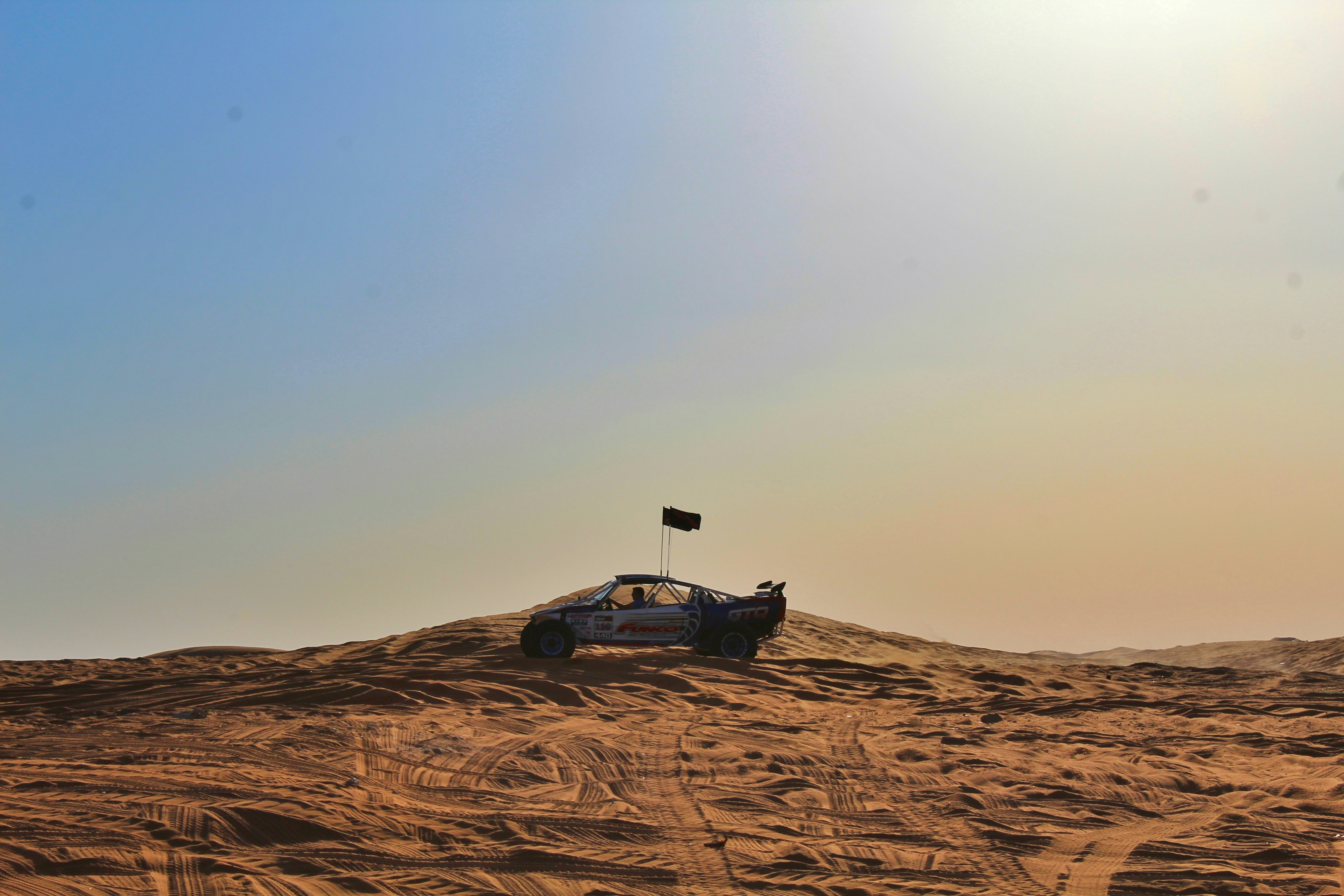 Red dune buggy cresting a massive sand dune, bright blue sky, deep shadows in desert valley below