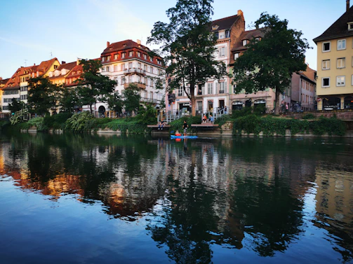 A picturesque riverside scene with charming historic buildings reflecting in the calm water. Trees and vibrant greenery line the riverbank, and people can be seen enjoying the serene atmosphere along the water's edge.