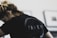 A person with light brown hair tied back is wearing a black shirt with the word 'INNER' printed on it in large letters. The focus is on the shirt, and the background is blurred, showing what appears to be framed pictures on a wall.