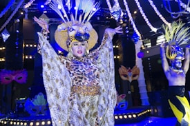A performer dressed in a lavish costume featuring a leopard print design and an elaborate headdress with horns is on stage. The setting is theatrical with bright stage lights and decorative elements like masks visible in the background. Another performer with a similar extravagant style is seen on the right, wearing a colorful headdress.