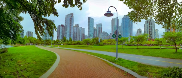 A city park featuring a paved walkway flanked by lush green grass and trees, with a background of modern high-rise buildings. A lamppost with signs for pedestrian and bicycle paths stands prominently. The setting is serene with a bright and clear sky above.