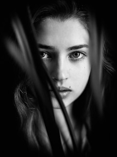A black and white portrait of a young person with an intense gaze, framed by blurred leaves or strands in the foreground. The composition creates a mysterious atmosphere, emphasizing the person's eyes and expression.