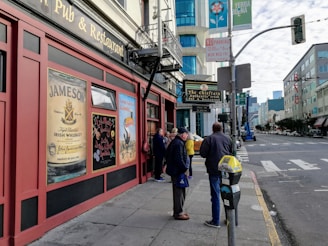 A street scene featuring a traditional Irish pub with posters advertising Jameson whiskey and Guinness. Several people are gathered in front of the pub, which is situated on a city sidewalk. The street is lined with modern buildings and traffic signs, with a partly cloudy sky above.