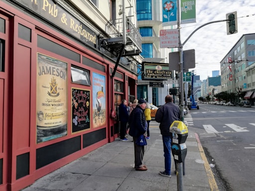A street scene featuring a traditional Irish pub with posters advertising Jameson whiskey and Guinness. Several people are gathered in front of the pub, which is situated on a city sidewalk. The street is lined with modern buildings and traffic signs, with a partly cloudy sky above.