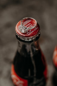 A close-up of a glass soda bottle with a red cap, topped with a silver ring featuring a large, round gemstone. The background is blurred, focusing attention on the bottle and the ring.