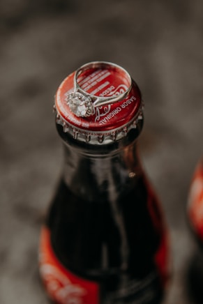 A close-up of a glass soda bottle with a red cap, topped with a silver ring featuring a large, round gemstone. The background is blurred, focusing attention on the bottle and the ring.