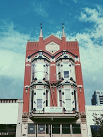 A historic red brick building with intricate architectural details, including pointed spires and arched windows. A 'For Lease' sign is prominently displayed on the building's facade. The sky is a bright blue with scattered clouds, providing a vivid background.