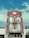 A historic red brick building with intricate architectural details, including pointed spires and arched windows. A 'For Lease' sign is prominently displayed on the building's facade. The sky is a bright blue with scattered clouds, providing a vivid background.