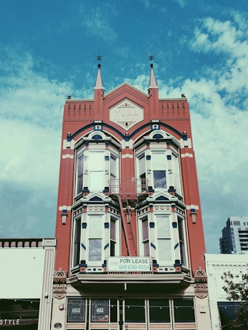 A historic red brick building with intricate architectural details, including pointed spires and arched windows. A 'For Lease' sign is prominently displayed on the building's facade. The sky is a bright blue with scattered clouds, providing a vivid background.