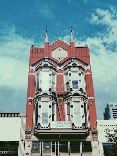 A historic red brick building with intricate architectural details, including pointed spires and arched windows. A 'For Lease' sign is prominently displayed on the building's facade. The sky is a bright blue with scattered clouds, providing a vivid background.