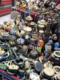 A collection of various antiques and brass ornaments displayed on a patterned cloth. The assortment includes intricate figurines, brass cups, a large key, padlocks, an hourglass, and decorative items like painted nesting dolls.