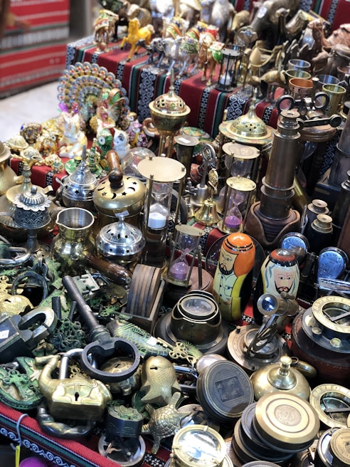 A collection of various antiques and brass ornaments displayed on a patterned cloth. The assortment includes intricate figurines, brass cups, a large key, padlocks, an hourglass, and decorative items like painted nesting dolls.