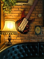 A cozy interior setting featuring a vintage-style lamp with a fringed shade, casting a warm glow against a textured brick wall. A decorative guitar with bold text is mounted on the wall alongside a few framed signs and lush green foliage hanging from above. A black, button-tufted leather bench provides seating below the display.