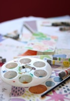 A tabletop covered with various art supplies including a circular paint palette with multiple paint colors, watercolor paint tubes, a paintbrush, and paper with colorful patterns and designs.
