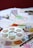 A tabletop covered with various art supplies including a circular paint palette with multiple paint colors, watercolor paint tubes, a paintbrush, and paper with colorful patterns and designs.