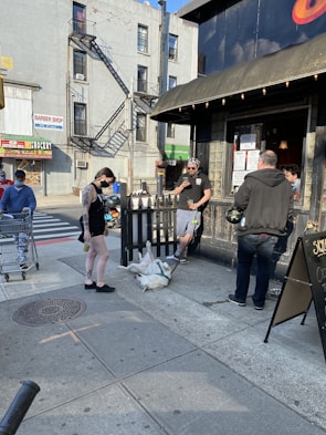 A group of people are gathered outside a building with one person walking a dog that is lying on its back on the sidewalk. There is a storefront in the background, a street with a cyclist and a person with a shopping cart on the side. The scene takes place in an urban setting with tall buildings and a fire escape visible on the building across the street.