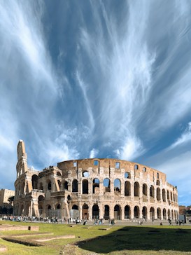 The image displays the ancient Colosseum in Rome under a dramatic sky with wispy clouds stretching above. A number of people can be seen walking around the historic site, highlighting its monumental scale.