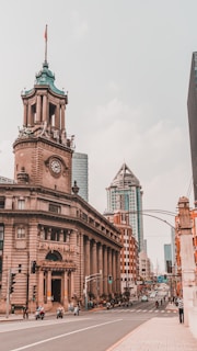 A city street features historic and modern architecture, with a prominent clock tower building adorned with a flag. The street is bustling with vehicles and pedestrians, and tall skyscrapers are visible in the background.