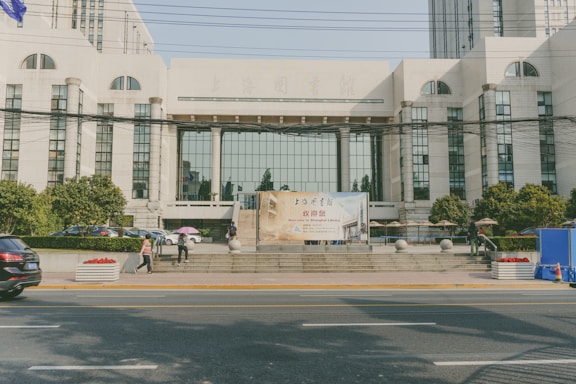 A large, modern building with tall windows and an impressive facade. In front, there is a banner with text in two languages welcoming visitors. People are walking nearby, and there are some cars parked outside. The setting includes greenery and is situated on a busy street with visible traffic and power lines overhead.