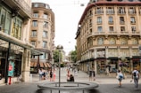 A European street scene with a mix of modern and historical architecture. The street is lined with ornate buildings, many featuring large windows and decorative facades. People are walking along the cobblestone path, and a modern art sculpture with a reflective sphere is centrally located. Trees and a carousel are visible in the background, adding to the urban atmosphere.