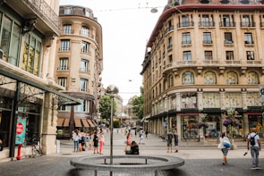 A European street scene with a mix of modern and historical architecture. The street is lined with ornate buildings, many featuring large windows and decorative facades. People are walking along the cobblestone path, and a modern art sculpture with a reflective sphere is centrally located. Trees and a carousel are visible in the background, adding to the urban atmosphere.