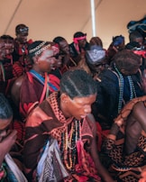 A group of people dressed in colorful, traditional clothing, adorned with beaded necklaces and headbands. They are gathered closely, sitting together in an indoor setting. The clothing features rich patterns and vibrant colors like red and brown, with some people wearing sunglasses or decorative hair accessories.