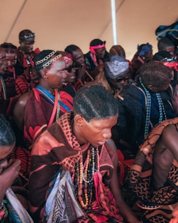 A group of people dressed in colorful, traditional clothing, adorned with beaded necklaces and headbands. They are gathered closely, sitting together in an indoor setting. The clothing features rich patterns and vibrant colors like red and brown, with some people wearing sunglasses or decorative hair accessories.
