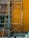 A construction scaffolding structure stands against a building facade with a brown and orange wall. The scaffolding is made of metal with visible wear and rust. A small bucket hangs from the structure, and scattered construction materials lie on the ground.