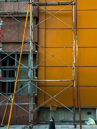 A construction scaffolding structure stands against a building facade with a brown and orange wall. The scaffolding is made of metal with visible wear and rust. A small bucket hangs from the structure, and scattered construction materials lie on the ground.