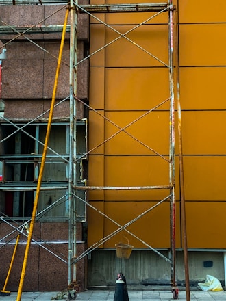 A construction scaffolding structure stands against a building facade with a brown and orange wall. The scaffolding is made of metal with visible wear and rust. A small bucket hangs from the structure, and scattered construction materials lie on the ground.