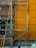 A construction scaffolding structure stands against a building facade with a brown and orange wall. The scaffolding is made of metal with visible wear and rust. A small bucket hangs from the structure, and scattered construction materials lie on the ground.