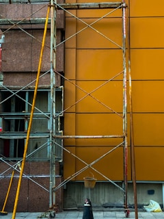 A construction scaffolding structure stands against a building facade with a brown and orange wall. The scaffolding is made of metal with visible wear and rust. A small bucket hangs from the structure, and scattered construction materials lie on the ground.