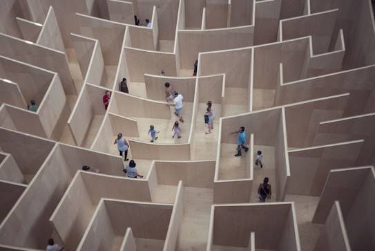 An indoor maze with high wooden walls is viewed from above, with several people, including children and adults, wandering through its pathways. The layout appears complex with numerous turns and corners.