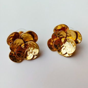 Two flower-shaped gold earrings with a textured surface set against a plain white background. The earrings have a shiny, reflective quality, displaying intricate details in their petal-like designs.