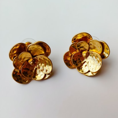 Two flower-shaped gold earrings with a textured surface set against a plain white background. The earrings have a shiny, reflective quality, displaying intricate details in their petal-like designs.