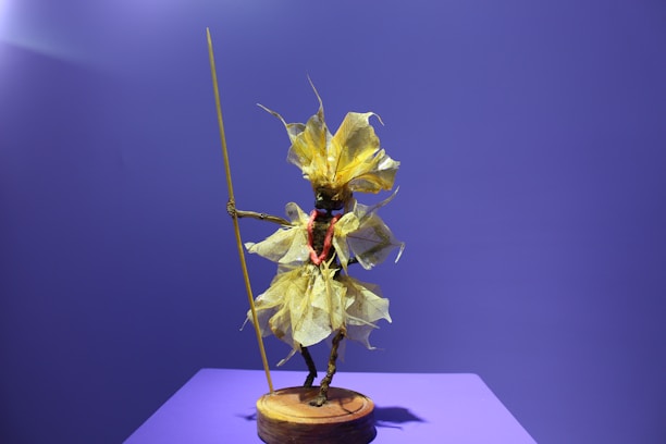 An artistic sculpture of a humanoid figure is crafted using leaves and natural materials, holding a long staff. The figure is mounted on a round wooden base against a plain purple background, highlighting its intricate details.