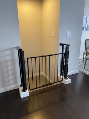 A black safety gate installed at the entrance of a stairway in an indoor space. The surrounding walls are painted a light neutral color, and the flooring is dark wood. There is a glimpse of a dining area with a patterned rug and a window in the background.