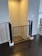 A black safety gate installed at the entrance of a stairway in an indoor space. The surrounding walls are painted a light neutral color, and the flooring is dark wood. There is a glimpse of a dining area with a patterned rug and a window in the background.