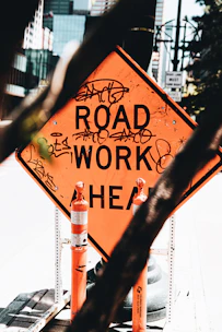 An orange diamond-shaped road work sign with graffiti is visible, surrounded by two traffic cones. The setting appears to be an urban area with blurred buildings and structures in the background. The sign is in the foreground, focusing attention with its vivid color amidst a muted backdrop.