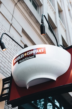 A large white bowl-shaped sign with the words 'Noodles & Company' and 'World Kitchen' is mounted outside a building. The sign is accompanied by black lights on both sides, and the building exterior features white brick and windows.