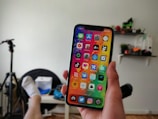 A person holding a smartphone displaying a colorful home screen with various app icons in a room with a chair, a tripod, and shelves in the background.
