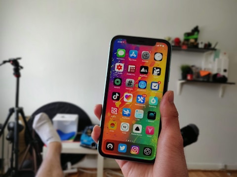 A person holding a smartphone displaying a colorful home screen with various app icons in a room with a chair, a tripod, and shelves in the background.