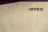 A textured wall with the word 'OFFICE' written in bold, dark lettering on the right side. The wall has an artistic pattern with faint shadows cast at an angle, adding depth and interest to the otherwise plain surface.