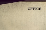 A textured wall with the word 'OFFICE' written in bold, dark lettering on the right side. The wall has an artistic pattern with faint shadows cast at an angle, adding depth and interest to the otherwise plain surface.