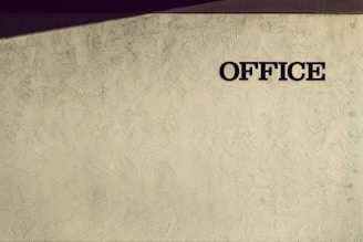 A textured wall with the word 'OFFICE' written in bold, dark lettering on the right side. The wall has an artistic pattern with faint shadows cast at an angle, adding depth and interest to the otherwise plain surface.