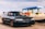 A black station wagon car is parked on a gravel surface near a container terminal. In the background, there are several colorful shipping containers stacked and a large blue and green gantry crane labeled 'INTERMOBIL'. The sky is partly cloudy.